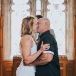 Elopement Wedding A couple shares a kiss during a wedding ceremony in front of a large, ornate stained-glass window. The woman, wearing a white lace dress, embraces the man, who is dressed in a black shirt. The wooden interior adds a warm ambiance to the intimate moment. Elopements Inc